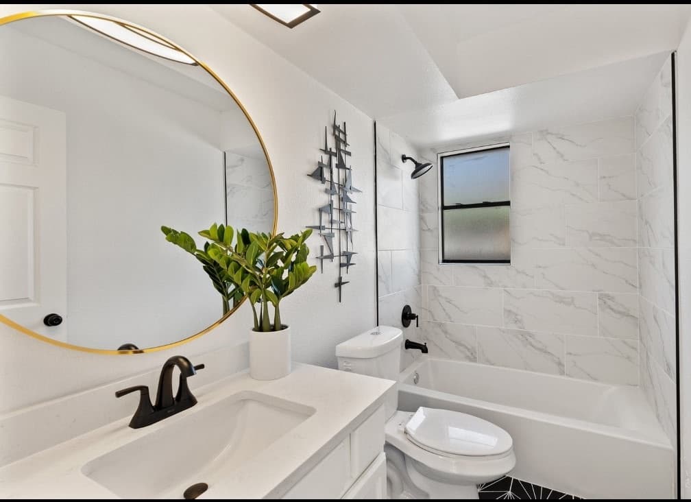 Bathroom remodel in Alamo Ranch San Antonio TX by Raging Bull Contracting — white marble tile tub surround with round gold-framed mirror and quartz vanity
