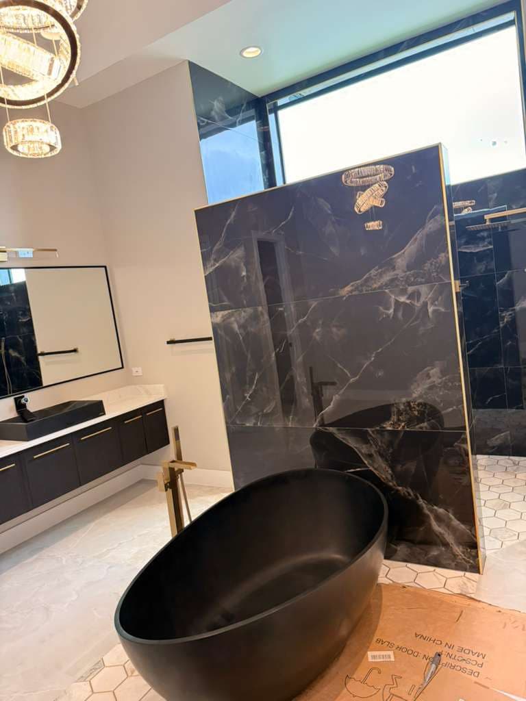 Bathroom remodel in Alamo Ranch San Antonio TX by Raging Bull Contracting — black marble-look slab shower walls with gold trim, freestanding matte-black tub and crystal chandeliers