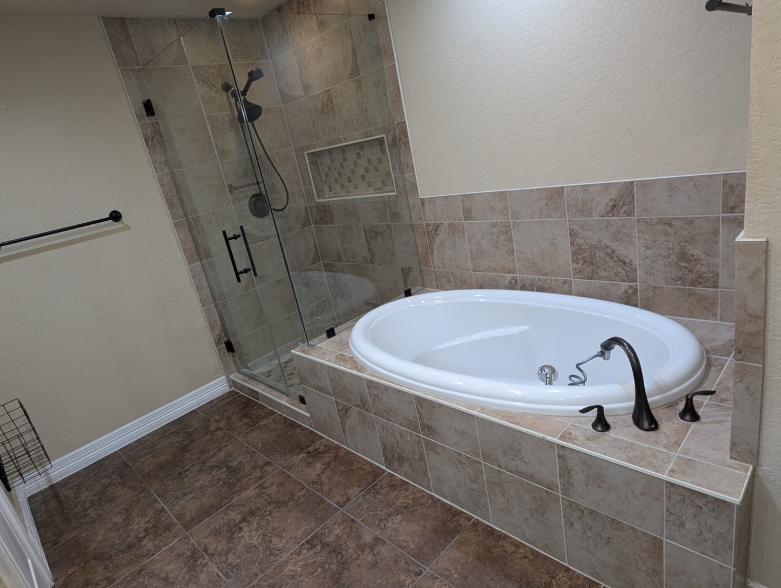 Walk-in shower rebuild in San Antonio TX by Raging Bull Contracting — frameless glass corner shower in travertine-look tile with a glass mosaic niche and oil-rubbed bronze fixtures, beside a tiled drop-in tub deck