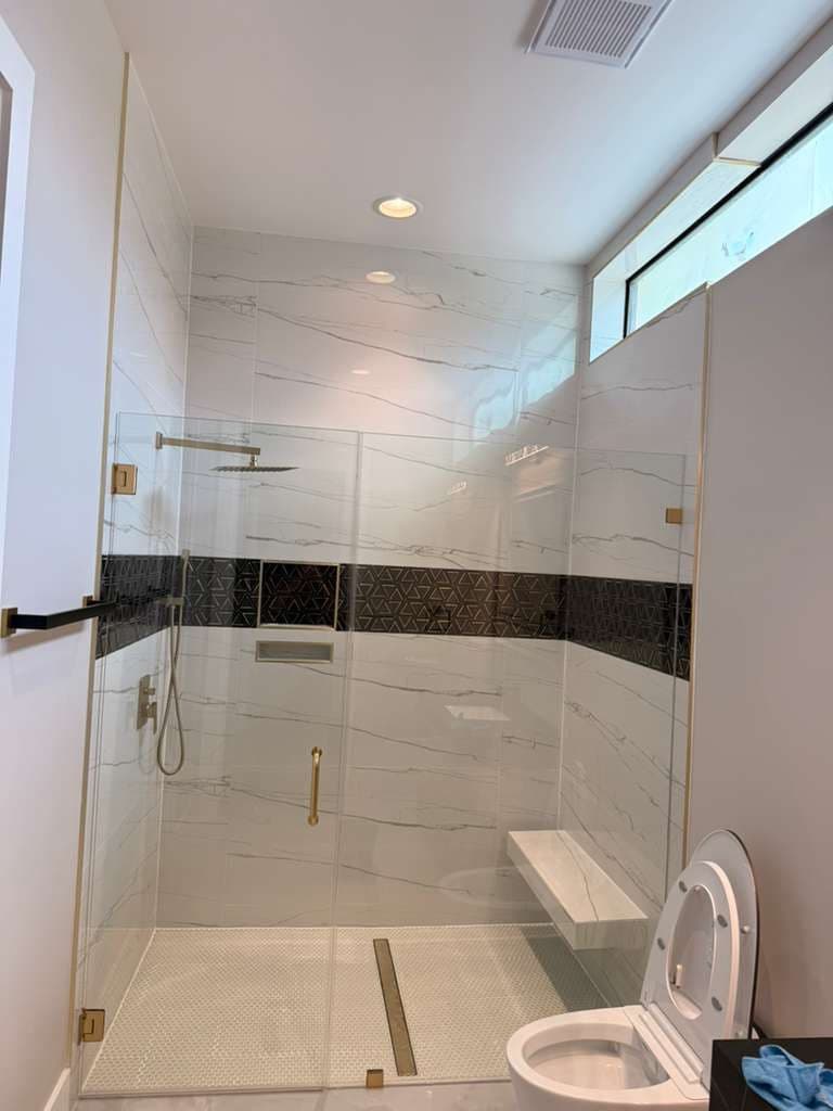 Walk-in shower remodel in Alamo Ranch San Antonio TX by Raging Bull Contracting — marble-look porcelain with black geometric mosaic accent band, brushed-gold fixtures, frameless glass and floating bench